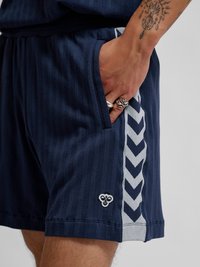 Ręka z pierścionkami spoczywająca w kieszeni granatowych szortów z jasnym wzorem jodełki i małym wyszywanym logo w pobliżu brzegu.