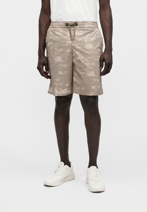 Άντρας που φοράει μπεζ σορτς με σχέδιο και κορδόνι, λευκά αθλητικά παπούτσια και λευκό πουκάμισο, στέκεται μπροστά σε απλό φωτεινό φόντο.