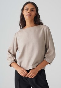 Beige Langarmbluse, glatte Textur, weit geschnittener U-Boot-Ausschnitt und geraffte Ärmel, kombiniert mit schwarzen Hosen.