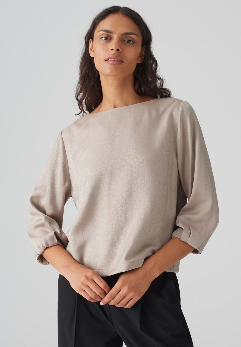 Beige Langarmbluse, glatte Textur, weit geschnittener U-Boot-Ausschnitt und geraffte Ärmel, kombiniert mit schwarzen Hosen.