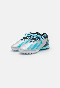 Las zapatillas de fútbol plateadas tienen rayas turquesa, cordones negros y una superficie texturizada. La suela cuenta con tacos blancos para mayor tracción.