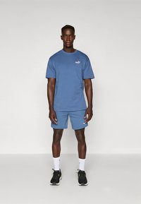 Sininen lyhythihainen t-paita ja siihen sopivat shortsit, jotka on valmistettu pehmeästä kankaasta. Rinnassa pieni valkoinen logo. Mustat lenkkitossut ja valkoiset sukat.