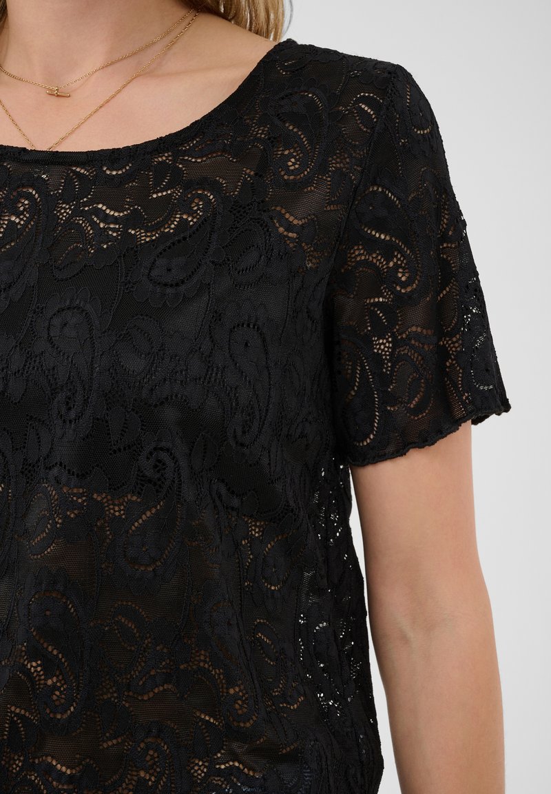 Haut noir en dentelle à manches courtes avec des motifs floraux et cachemire porté par une personne, montrant le décolleté et une partie du bras avec deux colliers en or.