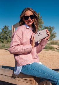 Žena v slunečních brýlích a růžovém svetru sedí na dřevěné lavici venku a drží světle šedou prošívanou peněženku s klipem.