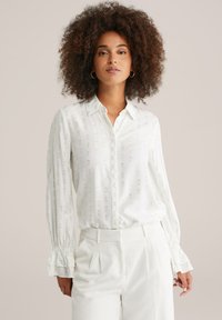 Camicia bianca con un delicato motivo a stampa e colletto a bottoni. Presenta maniche lunghe con polsini arricciati e una vestibilità comoda.