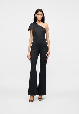 Woman wearing a fitted black one-shoulder jumpsuit with wide-leg pants and black open-toe heels, standing against a plain gray background.