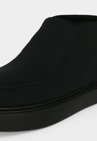 Μαύρο παπούτσι slip-on με λείο πάνω μέρος και παχιά σόλα, εμφανίζεται σε κοντινή γωνία σε απλό φωτεινό φόντο.