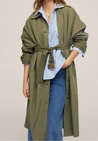 Femme portant un trench-coat olive ceinturé sur une chemise rayée bleu et blanc aux manches retroussées et un jean en denim bleu.