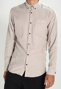 Beżowa koszula na długi rękaw zapinana na guziki, wykonana z lekkiego materiału, z kieszenią na piersi i ciemnymi guzikami jako akcentami.