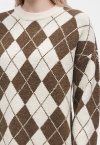 Pull en laine avec motif losange argyle marron et crème porté par une personne aux cheveux clairs et à la peau claire.