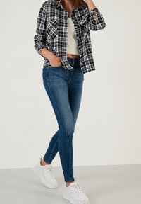 Camicia a quadri bianco e nero con due tasche sul petto, abbinata a jeans skinny blu scuro e sneakers bianche, esposta su uno sfondo semplice.