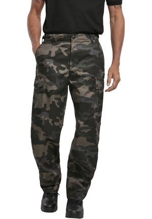 Hombre con camisa negra, botas negras y pantalones cargo de camuflaje verde oscuro y gris con bolsillos laterales, de pie frente a un fondo blanco.
