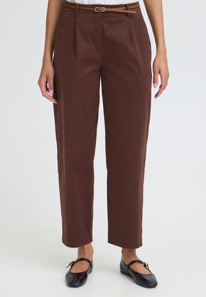 Kobieta ubrana w brązowe, wysokie spodnie o prostym kroju z zakładkami, wąski brązowy pasek oraz czarne baleriny typu Mary Jane na białym tle.