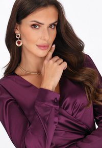 Blusa in raso di un profondo viola con scollo a V e polsini con bottoni. Il modello indossa orecchini a candelabro dorati e bordeaux, con una semplice collana attorcigliata.