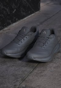 Pair of dark gray athletic sneakers with laces resting on a stone floor near a black wall.