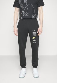 Pantalones deportivos negros con cintura elástica que presentan múltiples logos de Nike en blanco y amarillo en el lado izquierdo. Combinados con una camiseta gráfica negra y zapatos blancos.