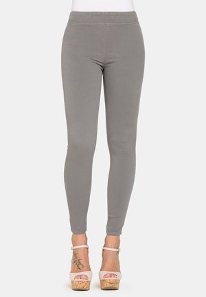 Donna con leggings aderenti grigi e sandali beige con zeppa e cinturini alla caviglia, in piedi su uno sfondo semplice.