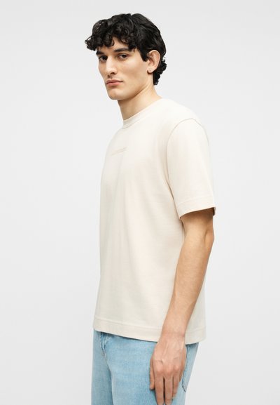 Νέος άνδρας με σγουρά σκουρόχρωμα μαλλιά, φοράει κοντομάνικο μπεζ πουκάμισο και ανοιχτόχρωμο μπλε τζιν, στέκεται πλευρικά μπροστά σε ένα απλό φόντο.