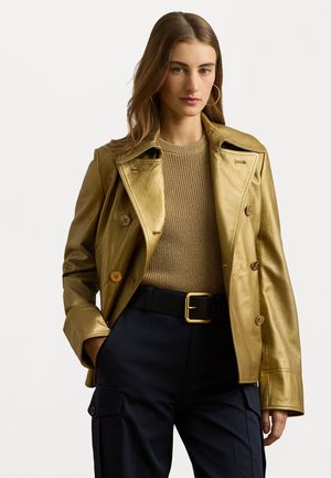 Guldmetallic læderjakke med dobbeltbryst og store knapper, båret over en ribbet beige sweatshirt og sorte bukser med bælte.