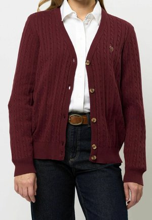 Femme portant une chemise blanche boutonnée sous un cardigan en maille torsadée bordeaux avec des boutons en bois, et un jean bleu foncé avec une ceinture marron.