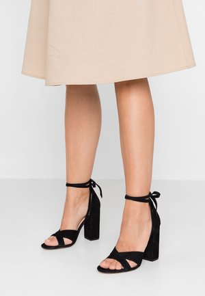 Sandales à talons hauts en daim noir avec bout ouvert, lien à la cheville et talon épais de forme carrée.