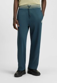 Teal jogger pants made of soft fabric with a drawstring waistband, featuring a striped accent and subtle logo detail on the side.