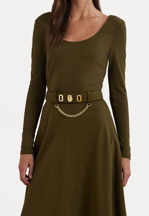 Femme portant une robe à manches longues vert olive avec une ceinture assortie ornée de détails dorés et une petite chaîne en or.