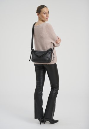 Naine, kes kannab beeži kampsunit ja musti nahkpükse, kannab õlal musta käekotti, vaadates üle õla.