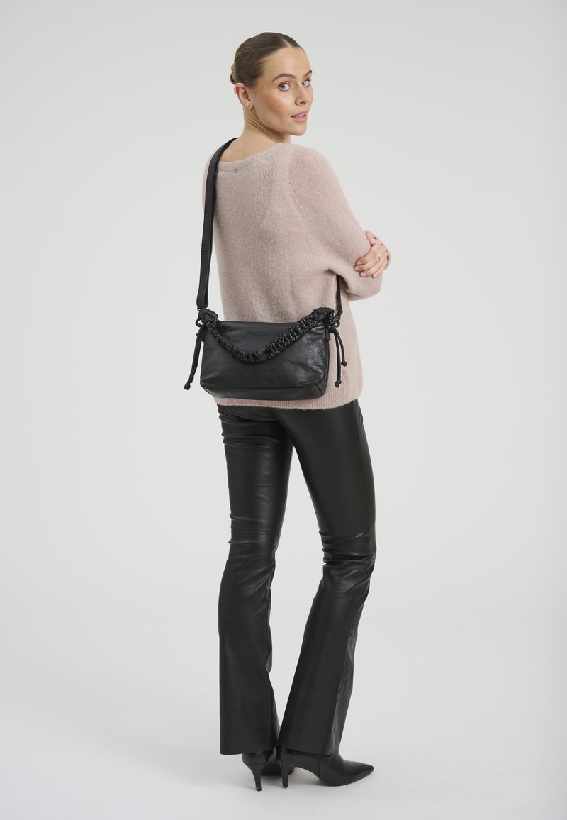 Femme portant un pull beige et un pantalon en cuir noir, avec un sac à main noir sur l'épaule, regardant par-dessus son épaule.