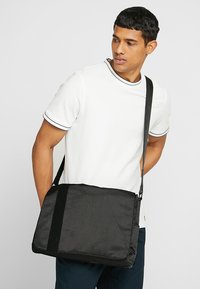 Svart budväska med diagonalt mönster, justerbar axelrem och slät textil. Har en klaffstängning och diskret logodetalj.