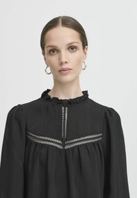 Blusa nera con scollo arricciato e maniche a sbuffo, caratterizzata da un dettaglio in rete sul petto. Tessuto morbido e strutturato con una vestibilità comoda.