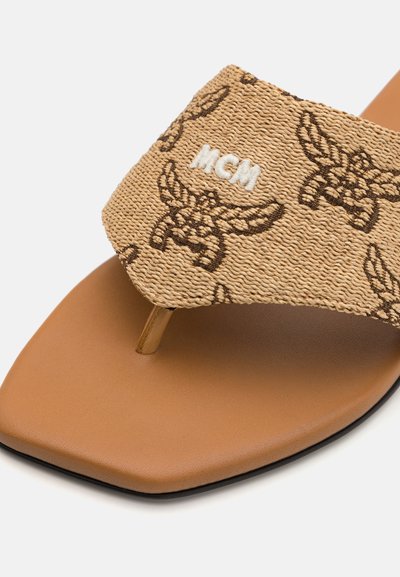 Des sandales en tan avec une bride en tissu texturé arborant un logo MCM marron et un design distinctif aux ailes, reposant sur une semelle marron.
