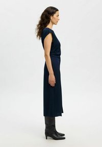 Femme aux longs cheveux ondulés se tient de profil, vêtue d'une robe midi sans manches bleu marine et de bottes noires à talons hauts arrivant au genou, sur un fond uni.