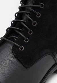 Gros plan sur une botte noire en cuir et daim avec des œillets métalliques noirs mats et des lacets noirs arrondis.