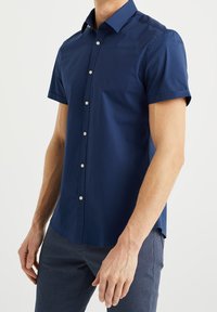 Muž v námořnicky modré košili s krátkým rukávem a tmavě modrých kalhotách, stojící s uvolněnýma rukama podél těla.