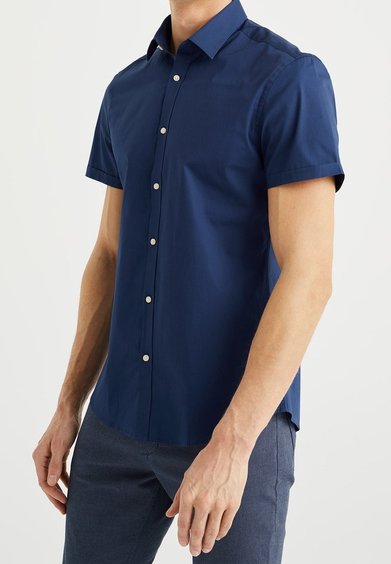 Muž v námořnicky modré košili s krátkým rukávem a tmavě modrých kalhotách, stojící s uvolněnýma rukama podél těla.