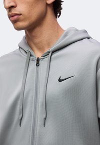 Gros plan sur une personne portant un sweat à capuche gris clair zippé avec cordons et un logo Nike swoosh noir sur la poitrine gauche.