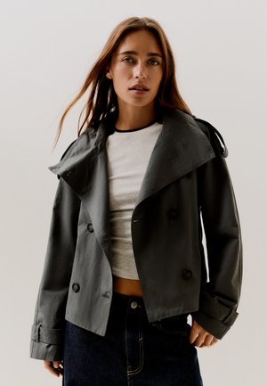 Giovane donna con lunghi capelli castani indossa una giacca doppiopetto grigio scuro sopra un top corto grigio chiaro e jeans blu scuro.