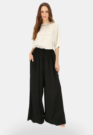 Kobieta stoi z długimi brązowymi włosami, ubrana w luźny biały krótki rękaw i szerokie czarne spodnie z rękami w kieszeniach.