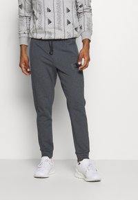 Dunkelgraue Jogginghosen mit elastischen Bündchen, Kordelzugbund und kleinem schwarzen Logo am linken Oberschenkel, kombiniert mit weißen Sportschuhen.