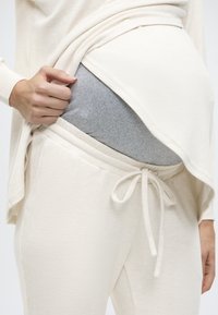 Μπεζ ανοιχτόχρωμο μακρυμάνικο τοπ φορεμένο πάνω από γκρι παντελόνι εγκυμοσύνης. Το τοπ έχει άνετη εφαρμογή με μαλακή υφή, και το παντελόνι διαθέτει ελαστική μέση με κορδόνι.