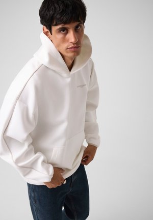 Giovane uomo con capelli scuri che indossa una felpa bianca con cappuccio e jeans blu, in piedi davanti a uno sfondo chiaro e uniforme.