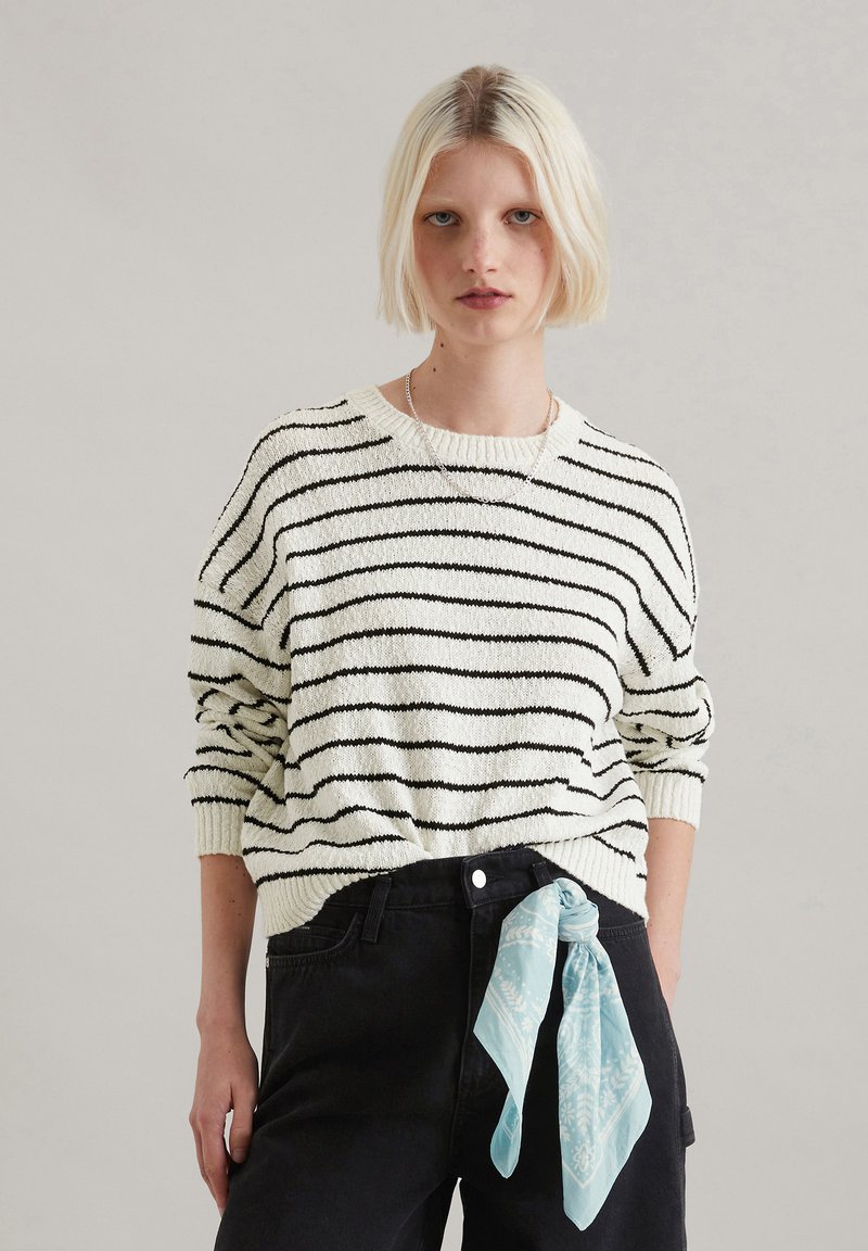 Cremfarbener Pullover mit schwarzen Streifen, geripptem Halsausschnitt und Bündchen. Kombiniert mit schwarzen Hosen und einem hellblauen, gemusterten Schal, der um die Taille gebunden ist.
