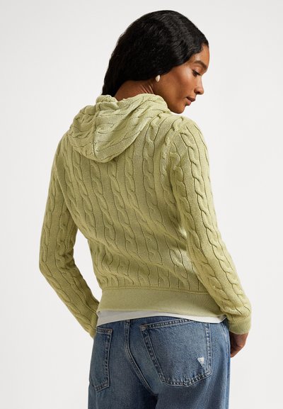 Moteris, dėvinti šviesiai žalią megztą džemperį su kapišonu ir mėlynas džinsus, matoma iš nugaros, žiūrinti per petį prieš paprastą foną.