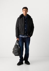 Black bomber jacket with faux fur collar, layered over a denim jacket, blue plaid shirt, dark jeans, and black shoes, holding a black tote bag.