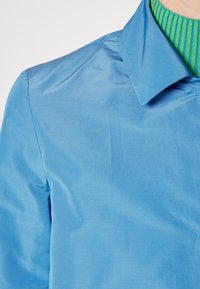 Camicia azzurro chiaro con una texture liscia e un colletto classico, indossata sopra un maglione verde a coste.
