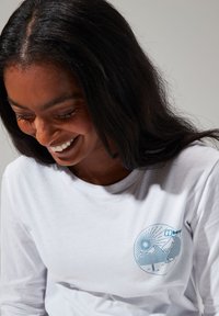 Ein weißes langärmliges T-Shirt mit einem blauen Grafikdesign von Bergen, Sonne und Bäumen im Brustbereich. Aus glattem Baumwollmaterial mit Rundhalsausschnitt.