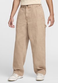 Person som bär rymliga beige byxor med stora frampockets och vita sneakers, stående mot en ljus, enfärgad bakgrund.