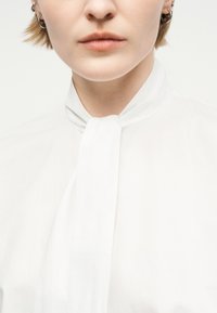 Κοντινό πλάνο ενός ατόμου που φοράει λευκό πουκάμισο με δεμένο μαντήλι στο λαιμό, εμφανίζοντας μέρος του προσώπου και κοντά μαλλιά με κρίκους σκουλαρίκια.