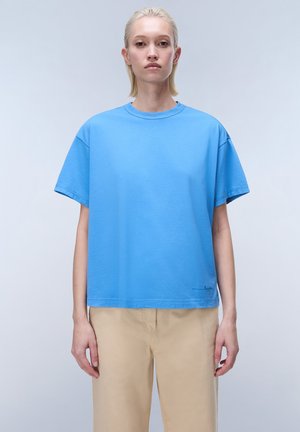 Jeune personne aux cheveux blonds courts portant un t-shirt ample bleu ciel et un pantalon beige, debout devant un fond clair uni.